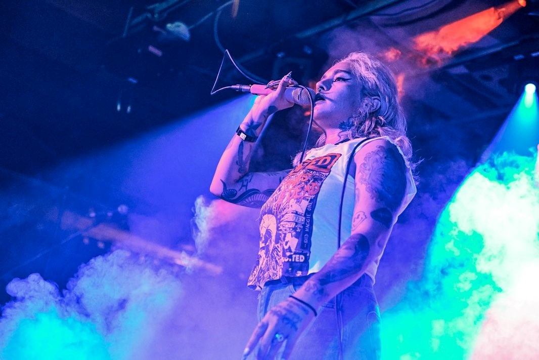 Tattooed singer holds microphone on smoky stage under blue and purple concert lights.