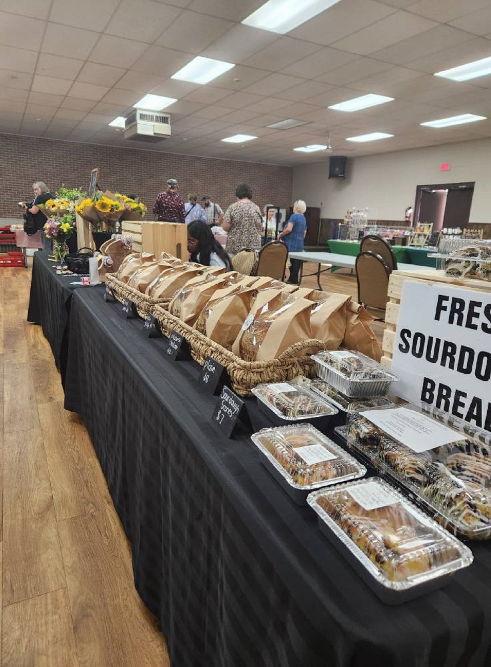 A market table with fresh sourdough bread in paper bags, baked goods in plastic containers, and bouquets of sunflowers on a black table, with people browsing in the background.