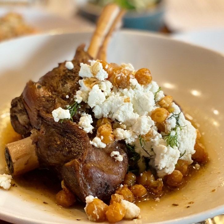 A savory lamb shank sits in a shallow white bowl, topped with crumbled feta cheese and golden chickpeas, creating a hearty and rustic gourmet dish.