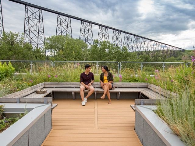 People on bench in lush park with steel bridge in background.