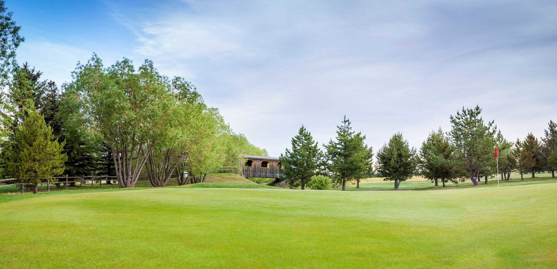 Golf green with red flag, trees and covered wooden bridge under blue sky.