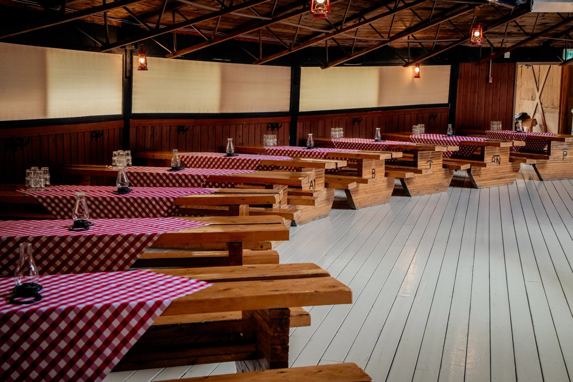 An empty dining hall with rows of wooden picnic tables covered in red and white checkered tablecloths, under a beamed ceiling with lanterns.