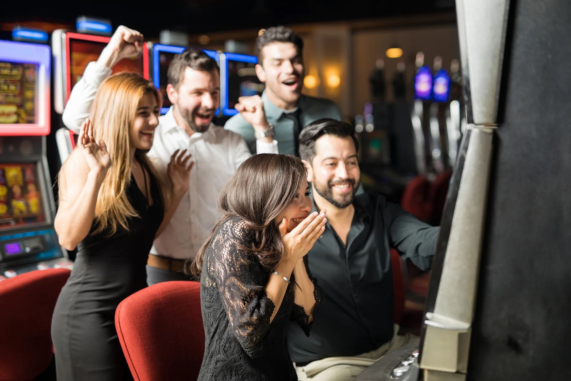Several casino guests gather around a slot machine as colorful gaming screens glow nearby.