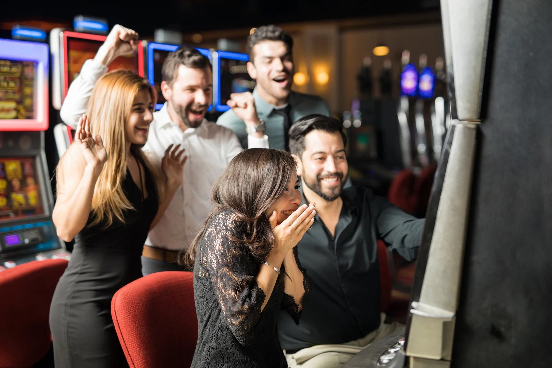 Several casino guests gather around a slot machine as colorful gaming screens glow nearby.