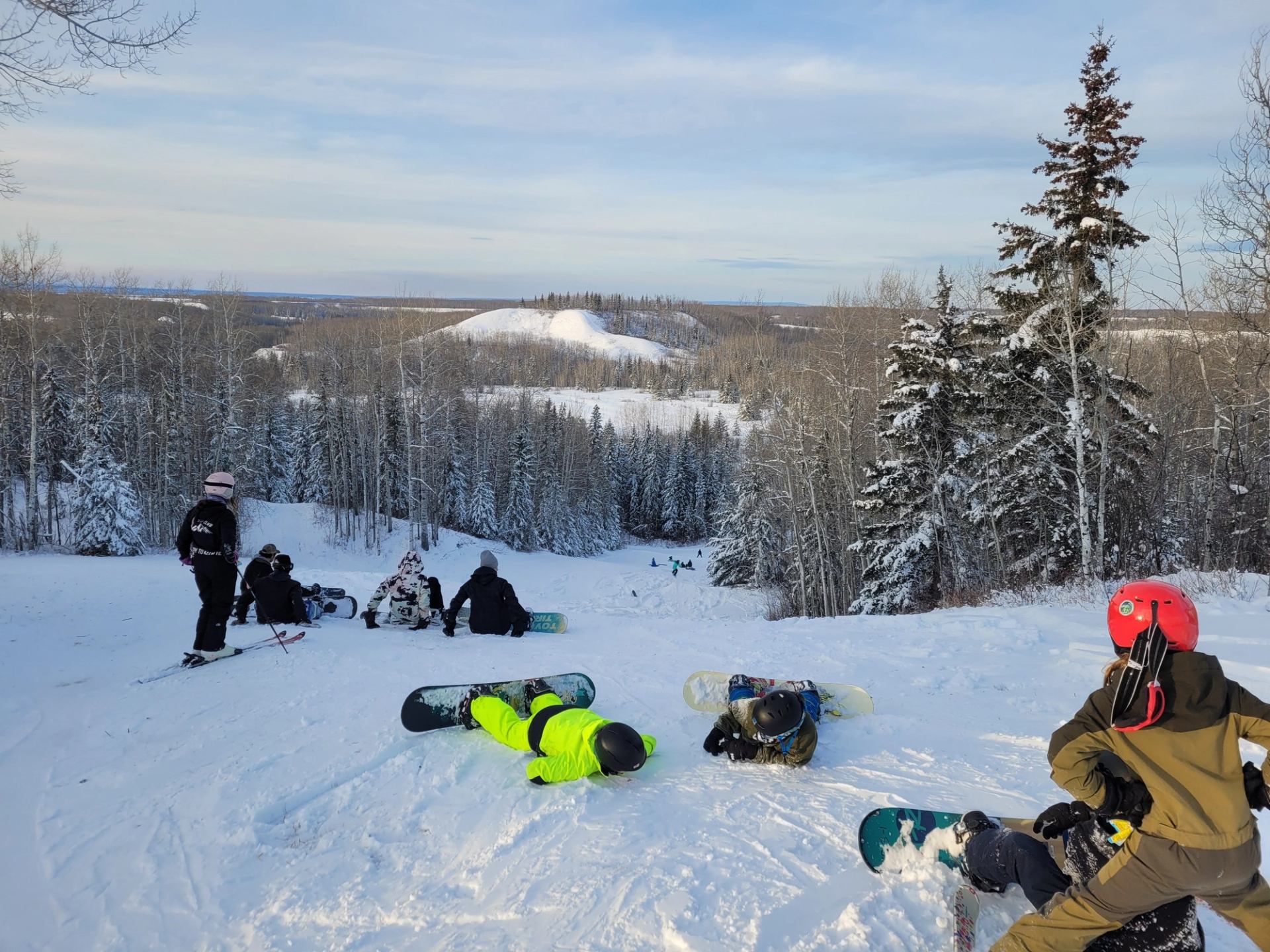 Skiers and snowboarders on a snowy hill with forest in the background.