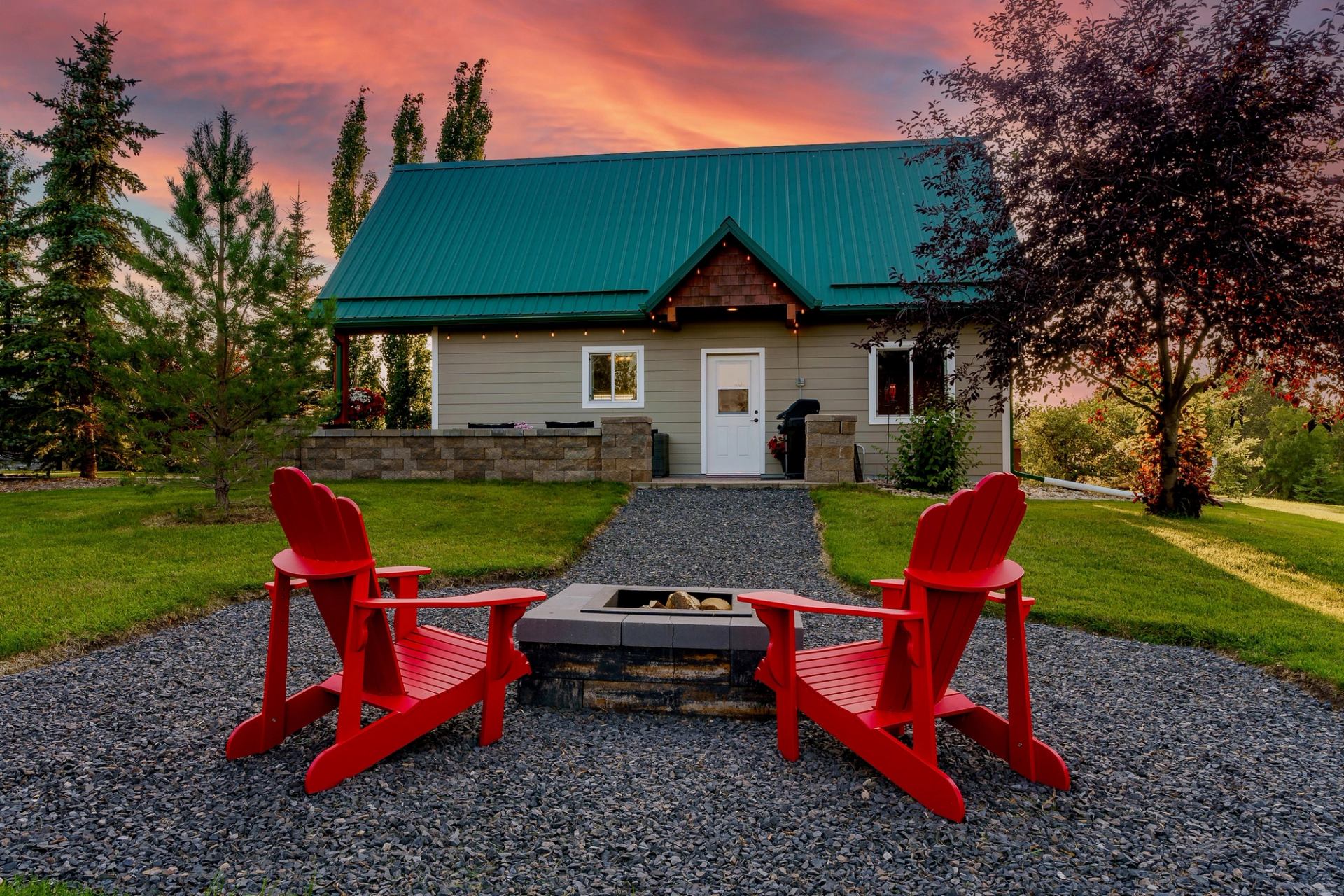 Two red Adirondack chairs around a firepit facing a cozy cabin with green roof at sunset.