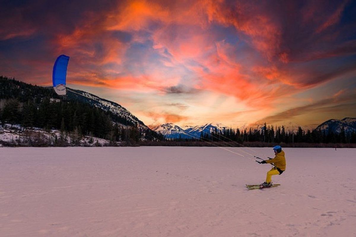 Person kite skiing across a snowy frozen lake at sunset, with mountains and a glowing sky in the background.