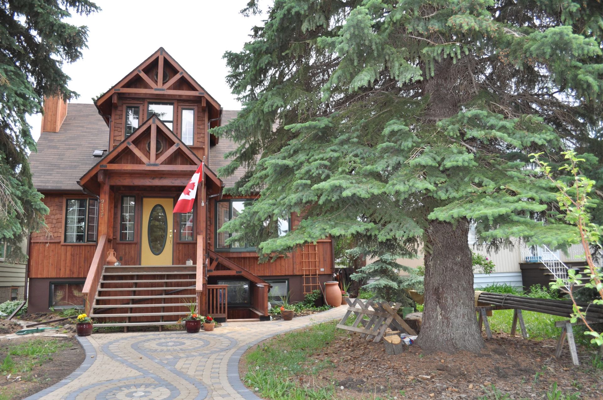 Wooden house with Canadian flag, stone walkway and large evergreen tree in front yard.