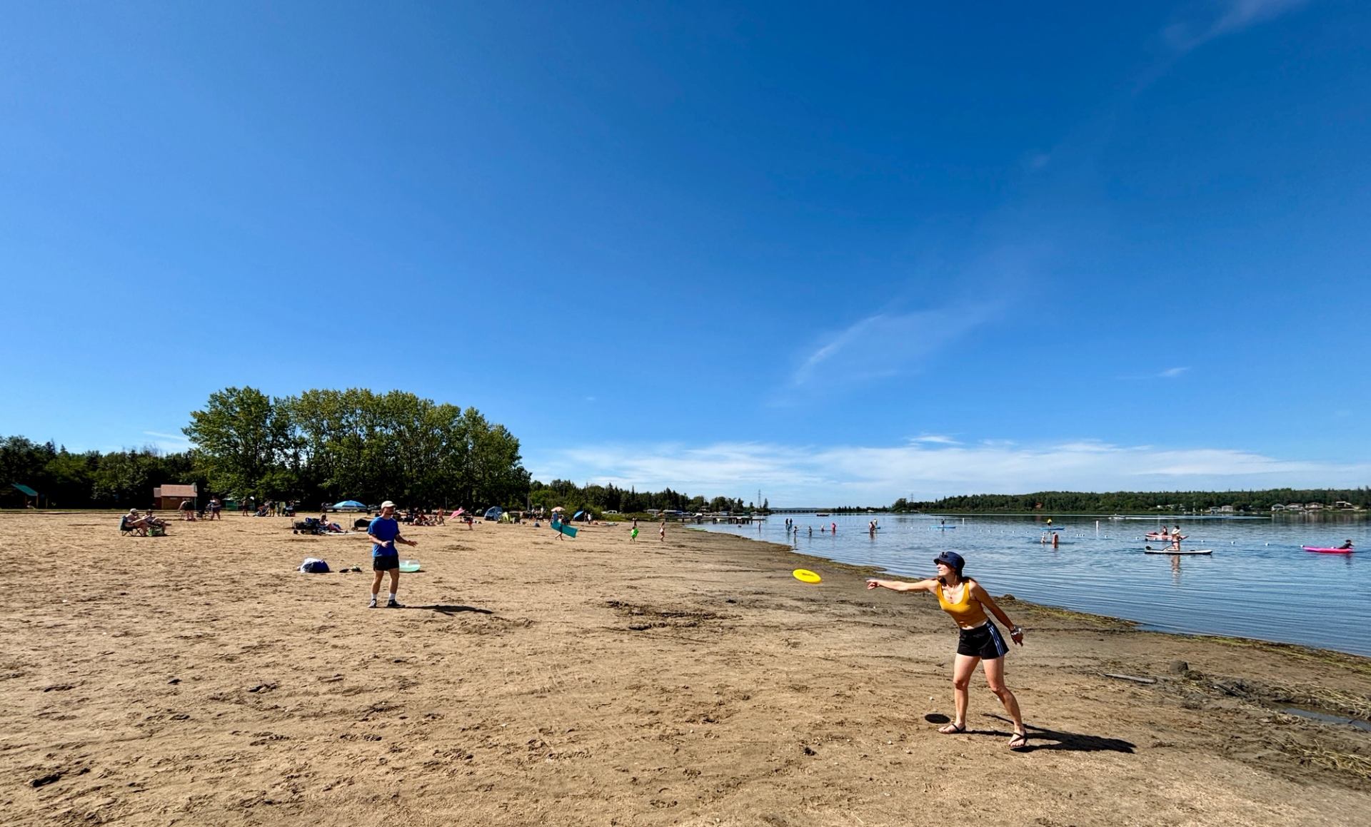 People play with a flying disc on a sandy beach beside a calm lake.