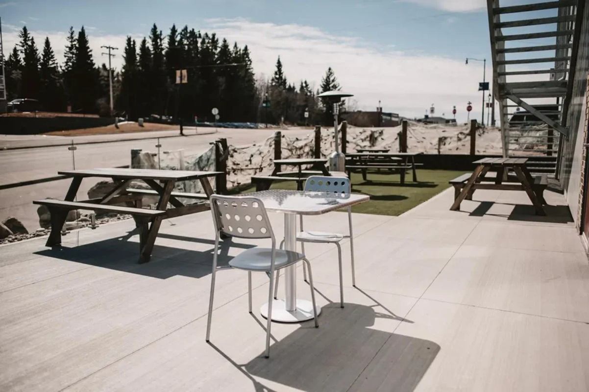 Outdoor patio with metal table, picnic benches, roadside view and trees in background.