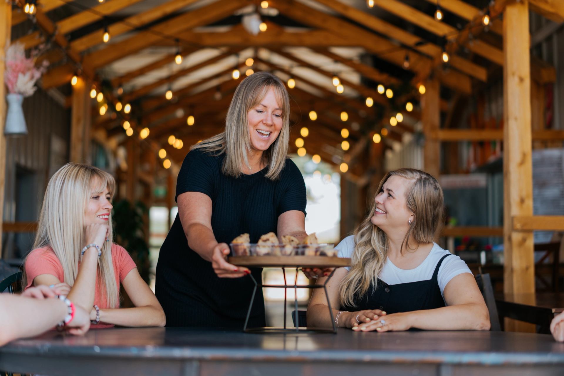 Cafe staff serving freshly baked treats to guests seated at a long table under warm string lights.