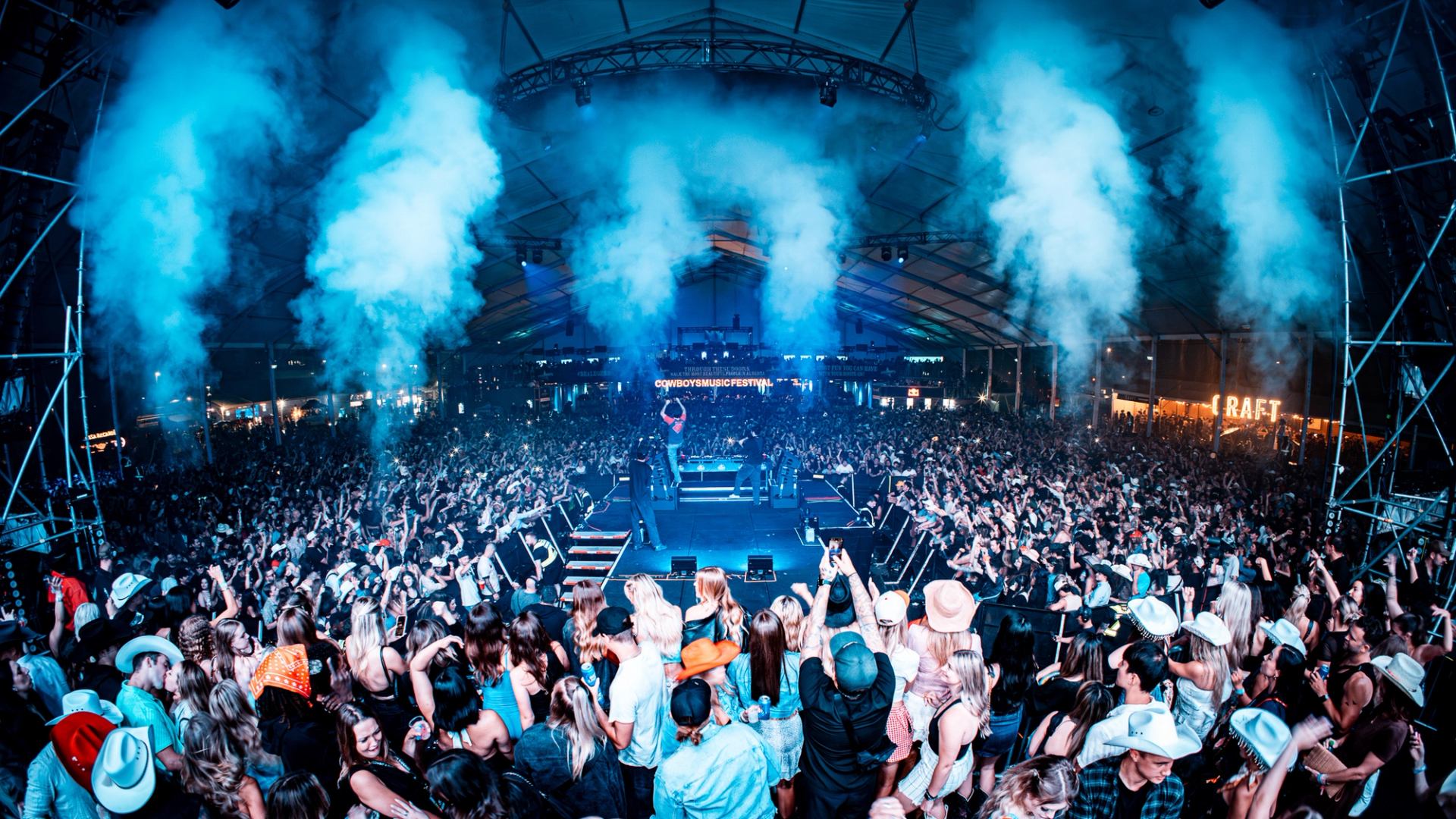 Massive festival crowd with smoke columns rising around the stage under a large tent.
