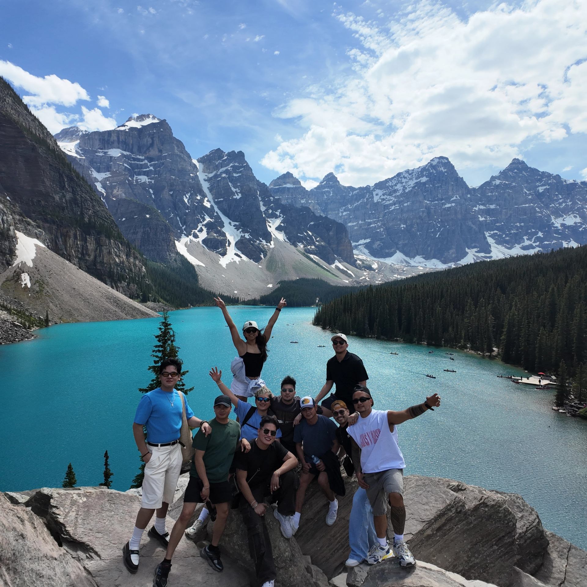 Group of people posing on rocky viewpoint above turquoise lake and snowy mountain peaks.