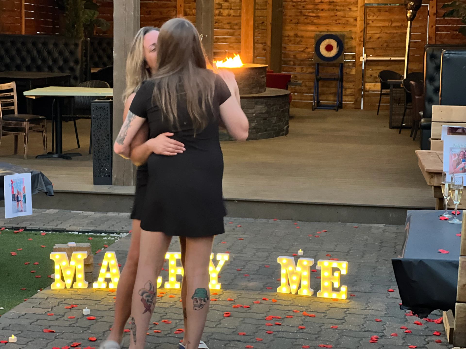 Outdoor proposal scene with rose petals, “Marry Me” lights, and a cozy fire pit.