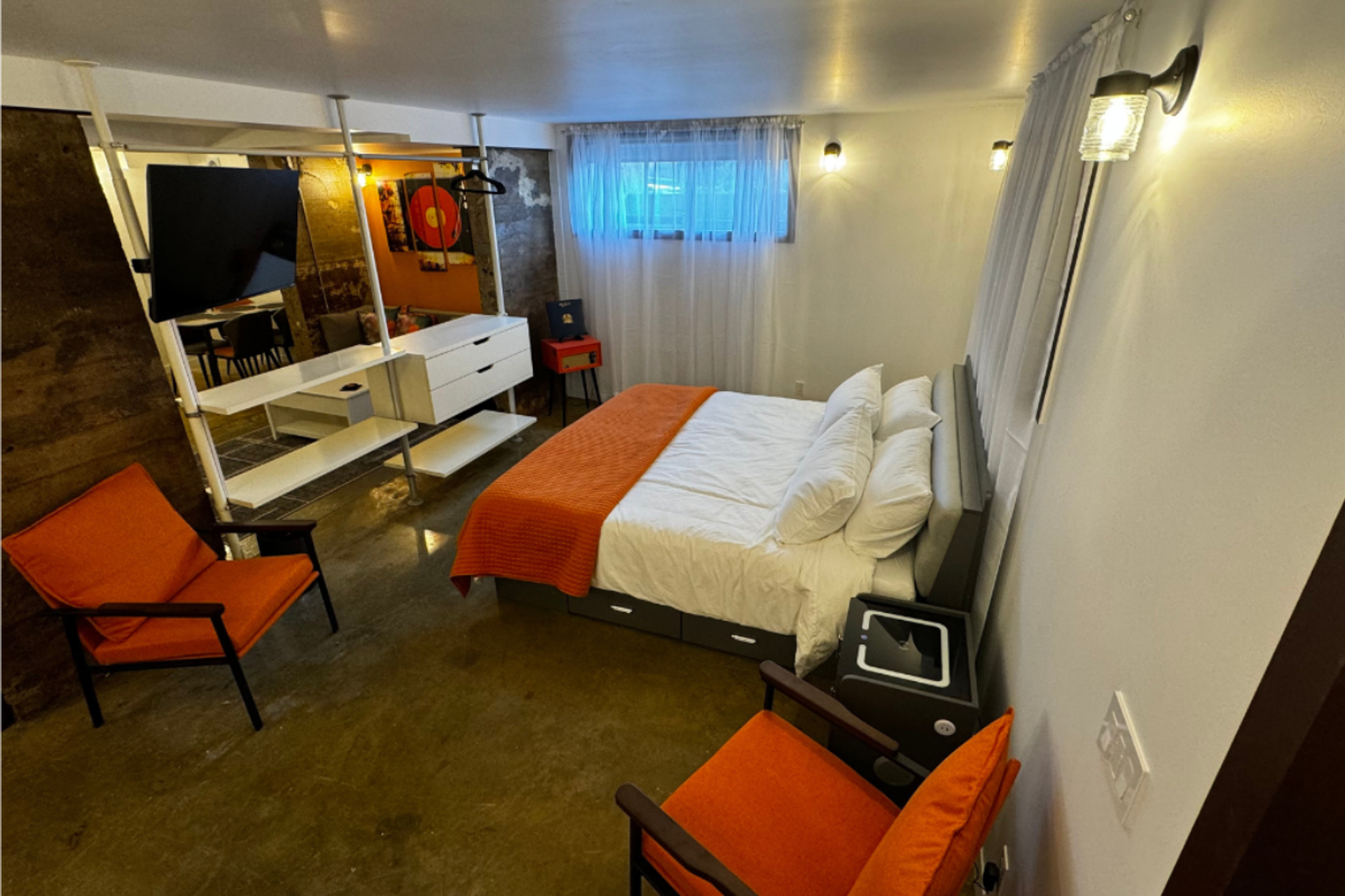 Bedroom with orange bedding, two orange chairs, wall-mounted TV and shelving unit.