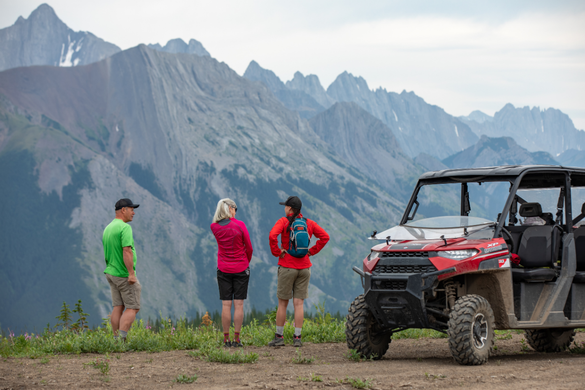 Three hikers stand beside a red off-road vehicle overlooking rugged mountain peaks.