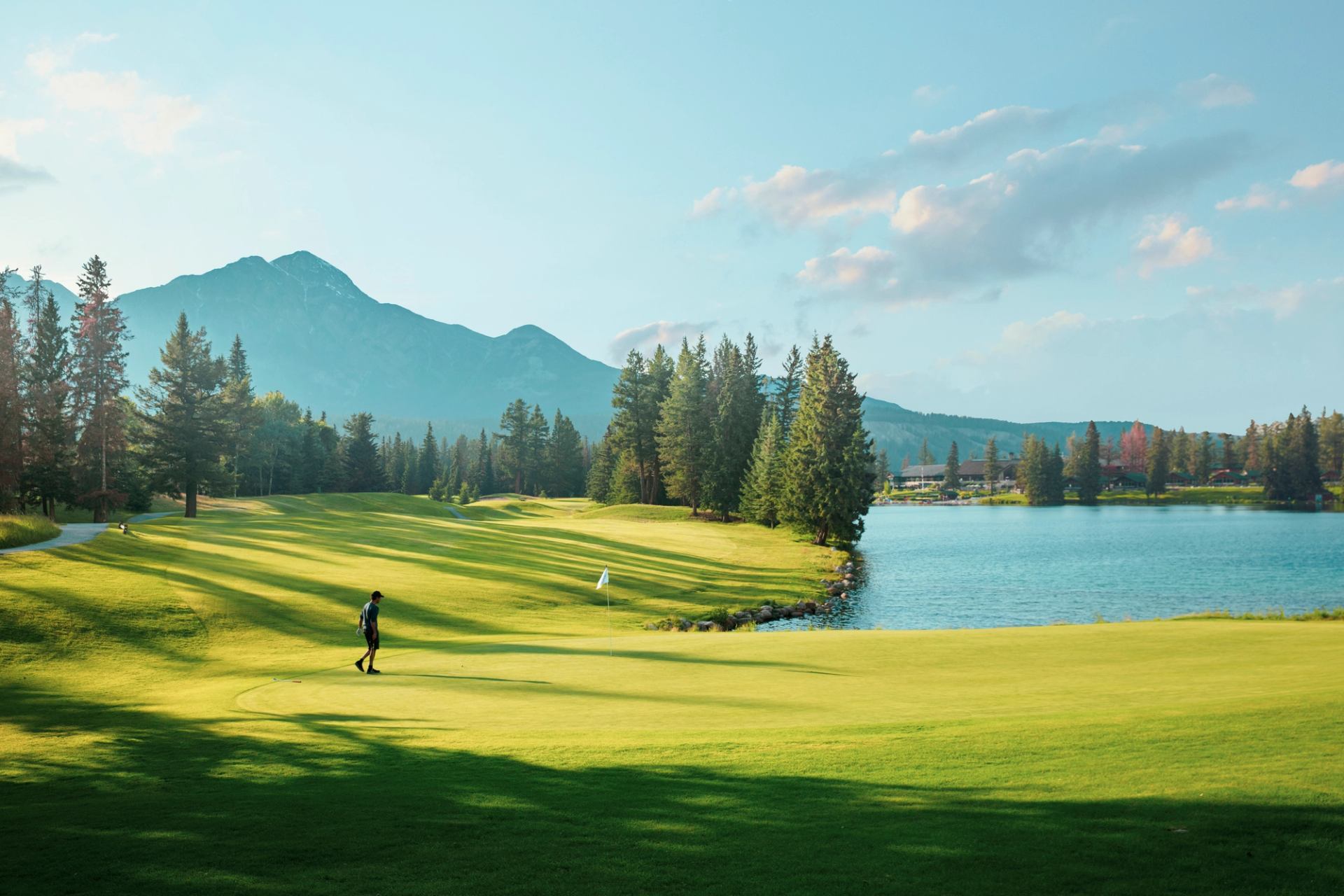 Golfer walks across green beside lake, with pine trees and mountains in background.