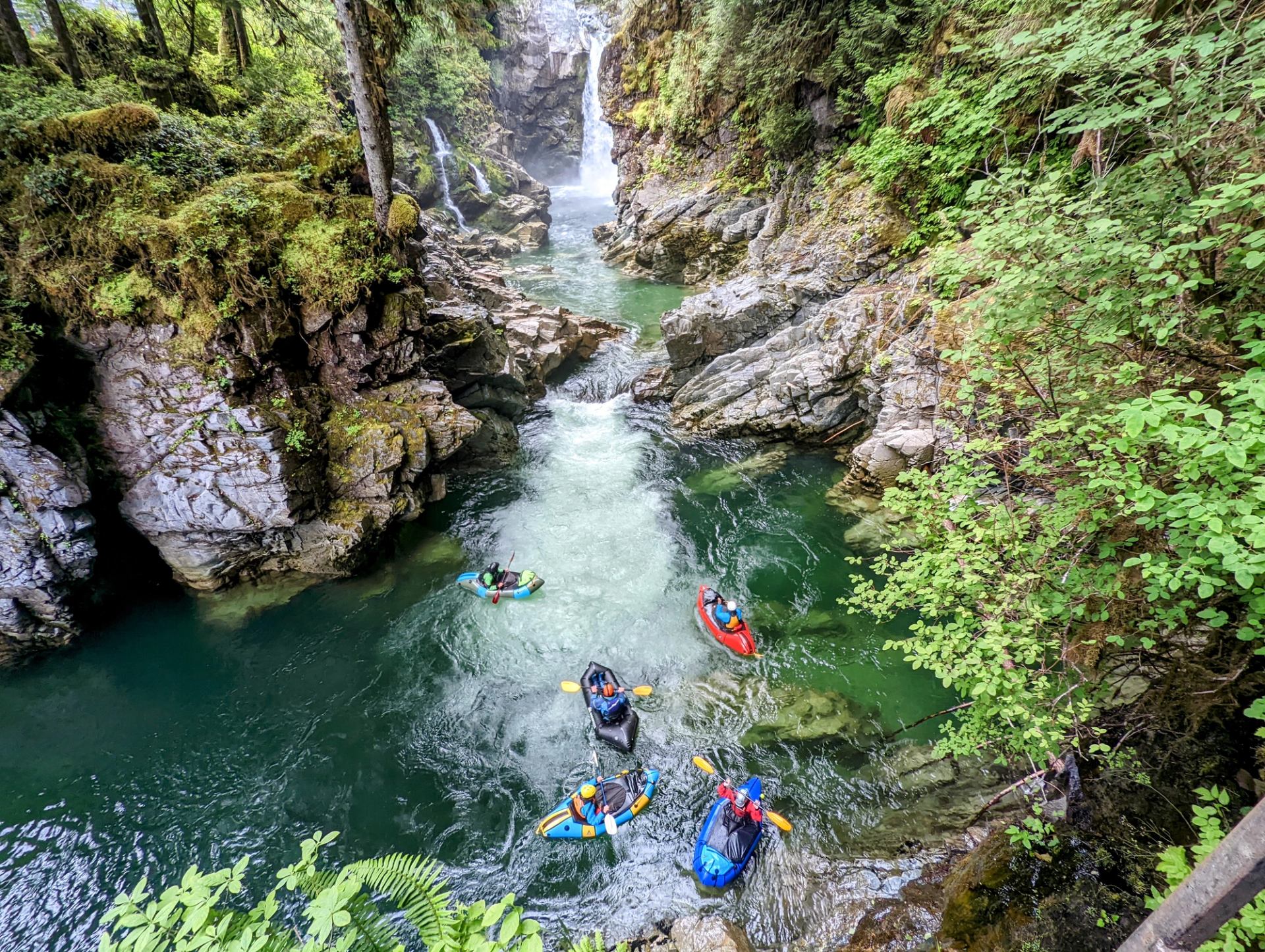 Kayakers paddle through narrow green canyon river beneath waterfalls and steep mossy cliffs.