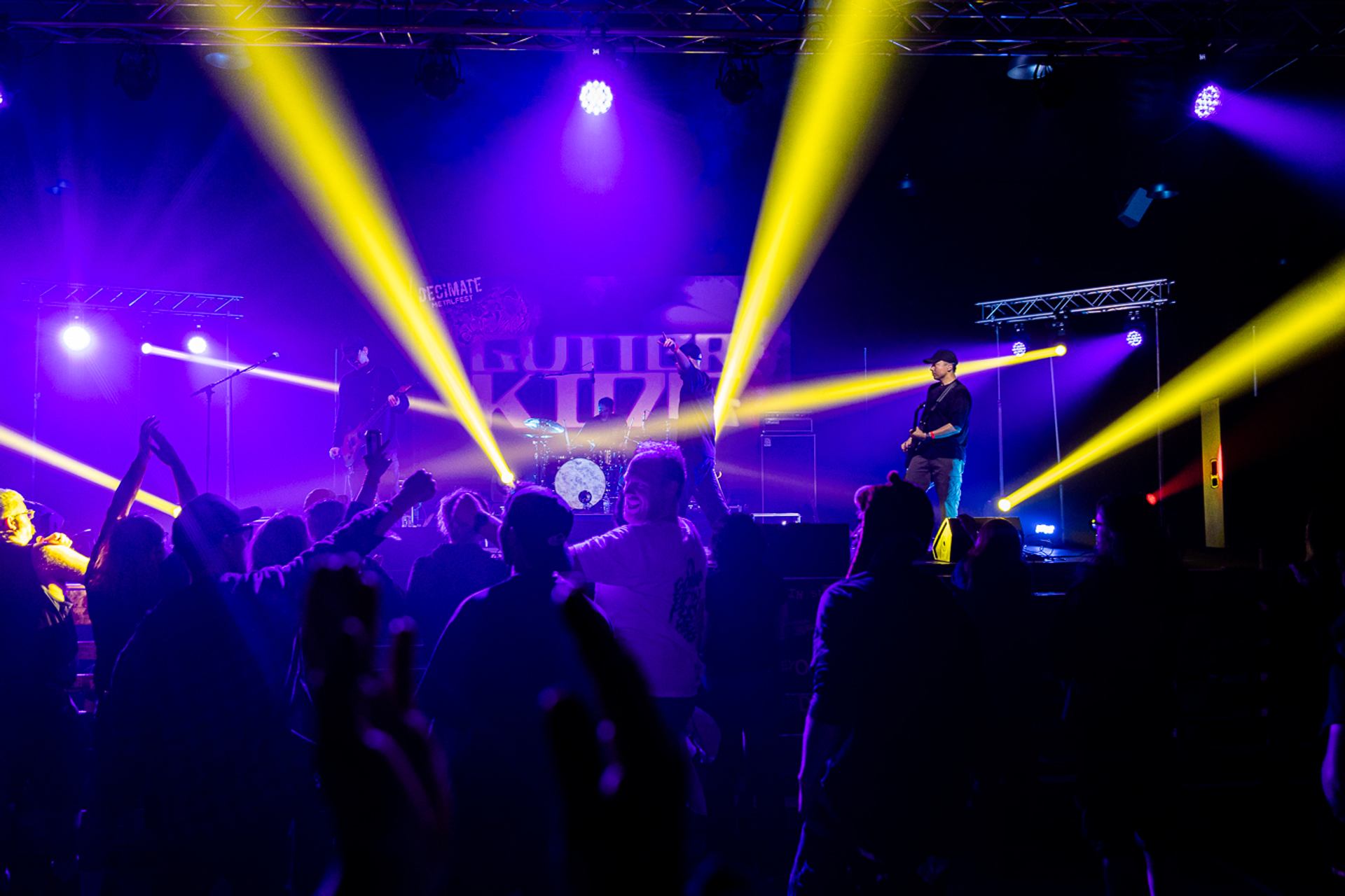 Decimate Metalfest 2026 shows a live band on stage with bright yellow beams and a crowd gathered in front under purple lighting.