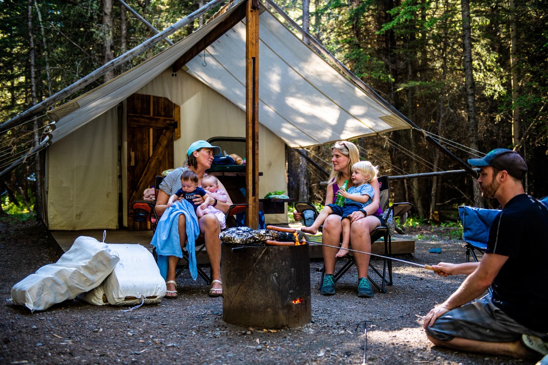 Campers sit by a fire pit outside a canvas tent in a forested campsite.