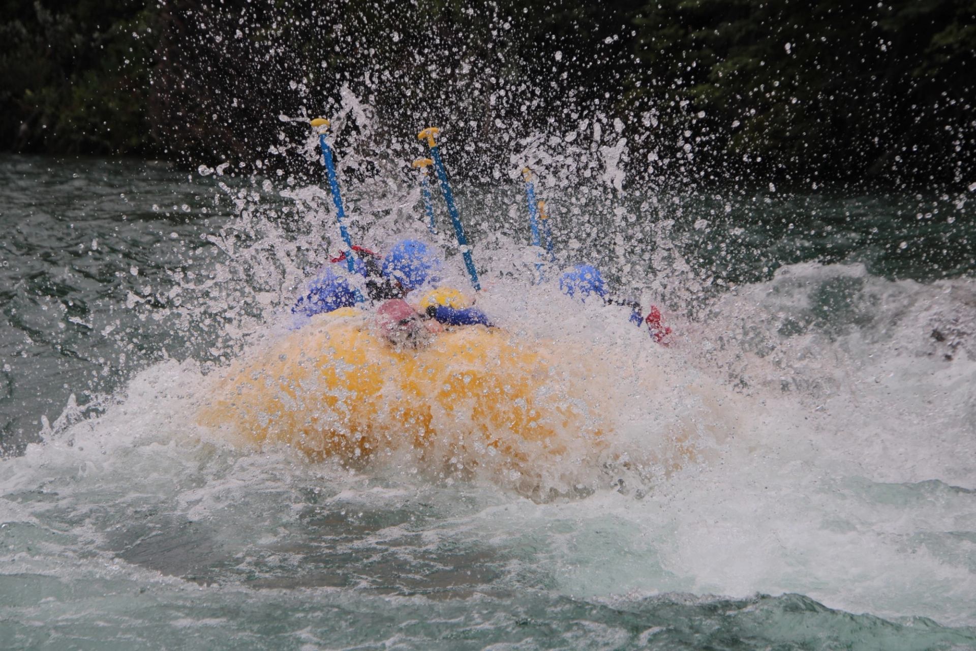 Yellow inflatable raft hits rapid, paddles and helmets partly hidden by splashing water.