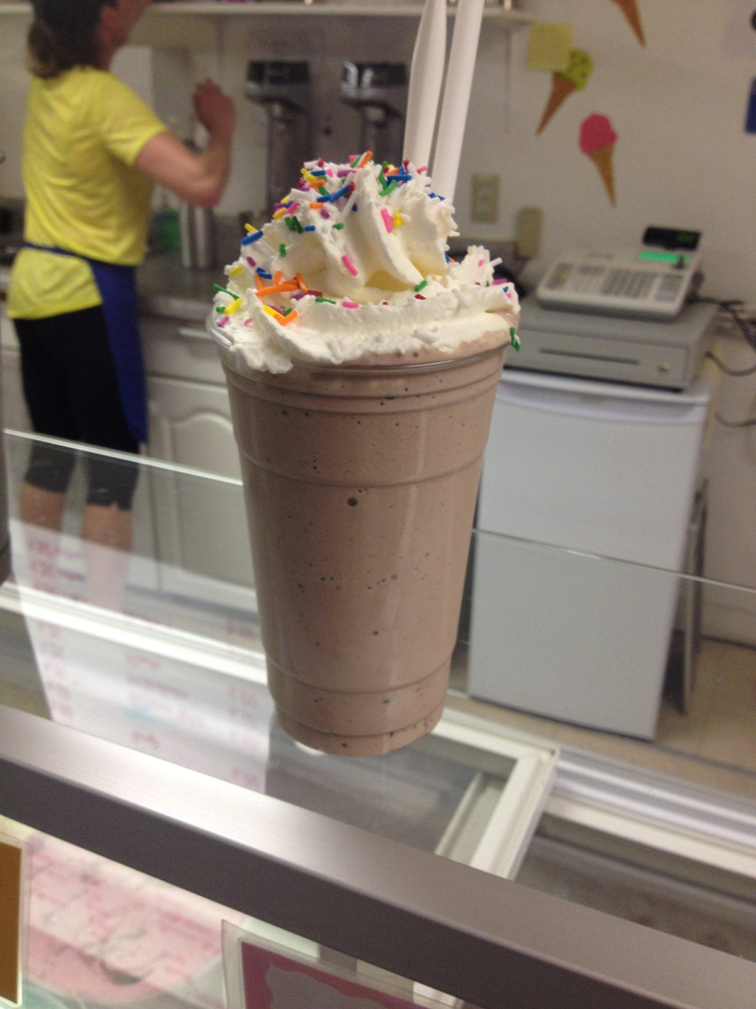 Chocolate milkshake topped with whipped cream and colorful sprinkles in a clear cup.