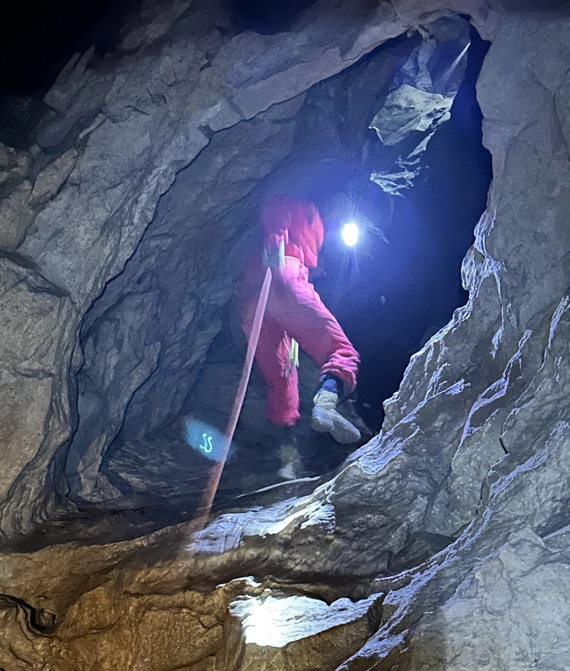 Person in red coveralls climbs through a narrow cave passage with a headlamp.