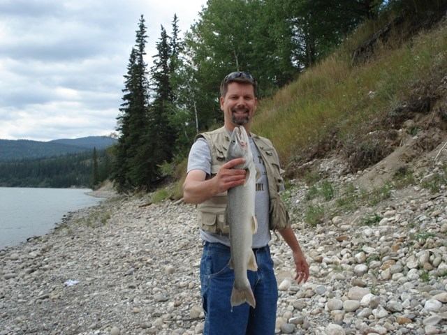 A smiling man holds a fish on a rocky shore by water, with forested hills in the background.