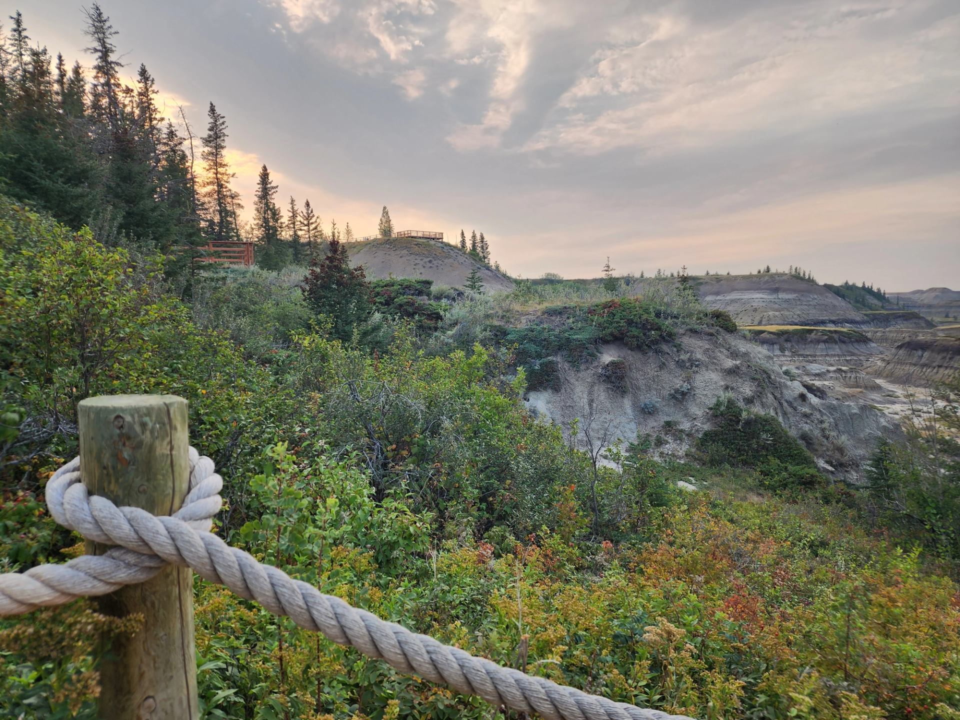  Scenic view of a lush green valley with mixed forest and eroded hills under a cloudy sunset sky, featuring a wooden post with a rope fence in the foreground.