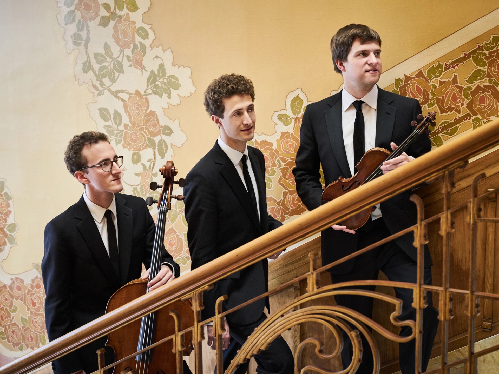 String musicians in suits walk up ornate staircase with cello and violin.