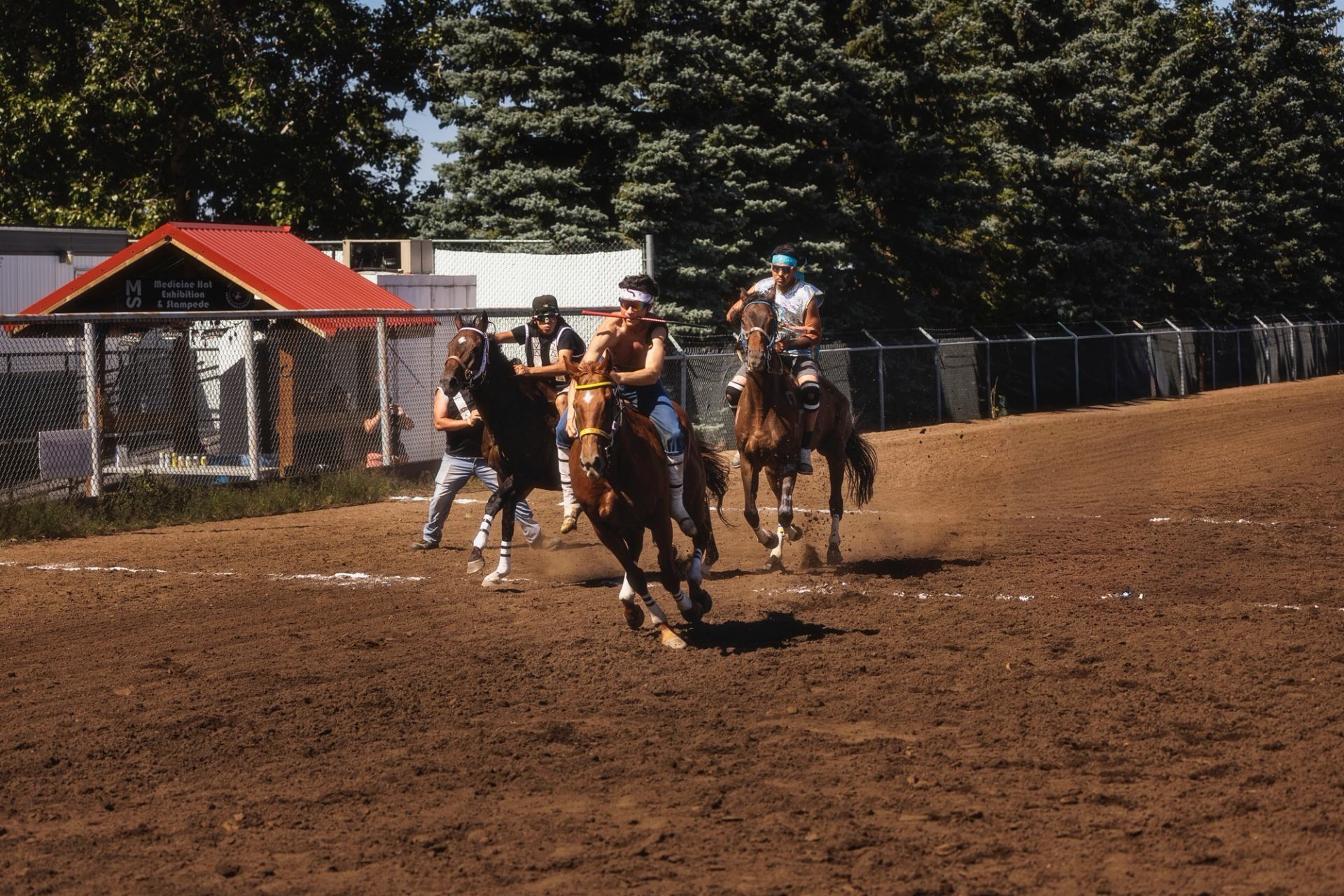 Bareback riders race horses on a dirt track beside a fence and trees.