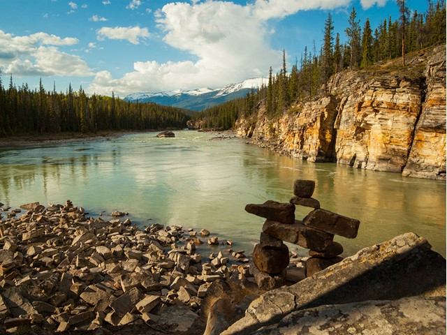 Rock inukshuk overlooks wide river between forested cliffs and distant snow-capped mountains.