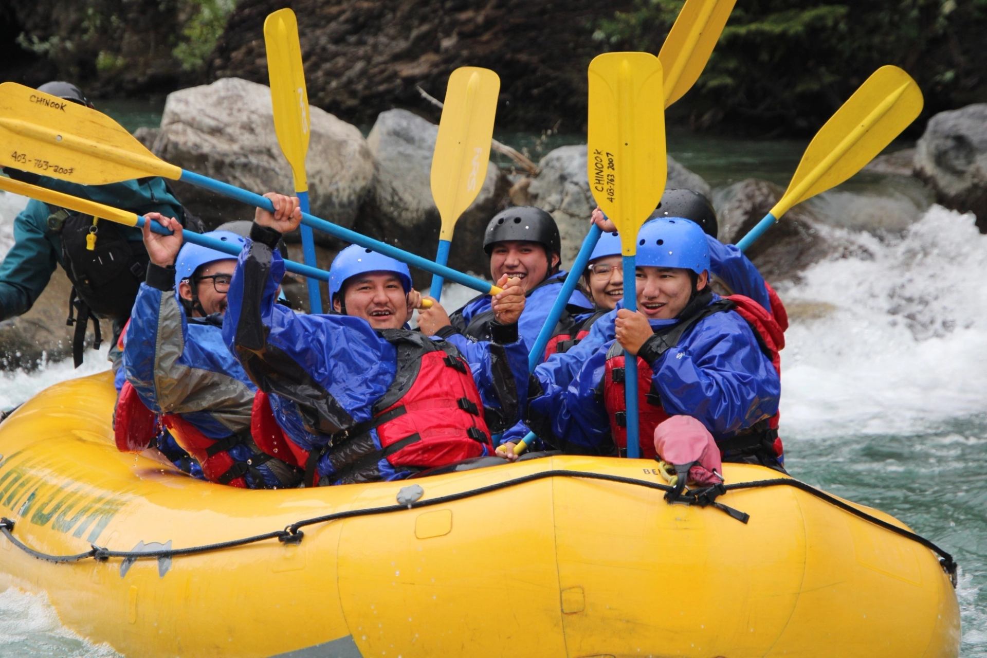 Group in helmets and lifejackets rafting in yellow inflatable boat, holding paddles upright.