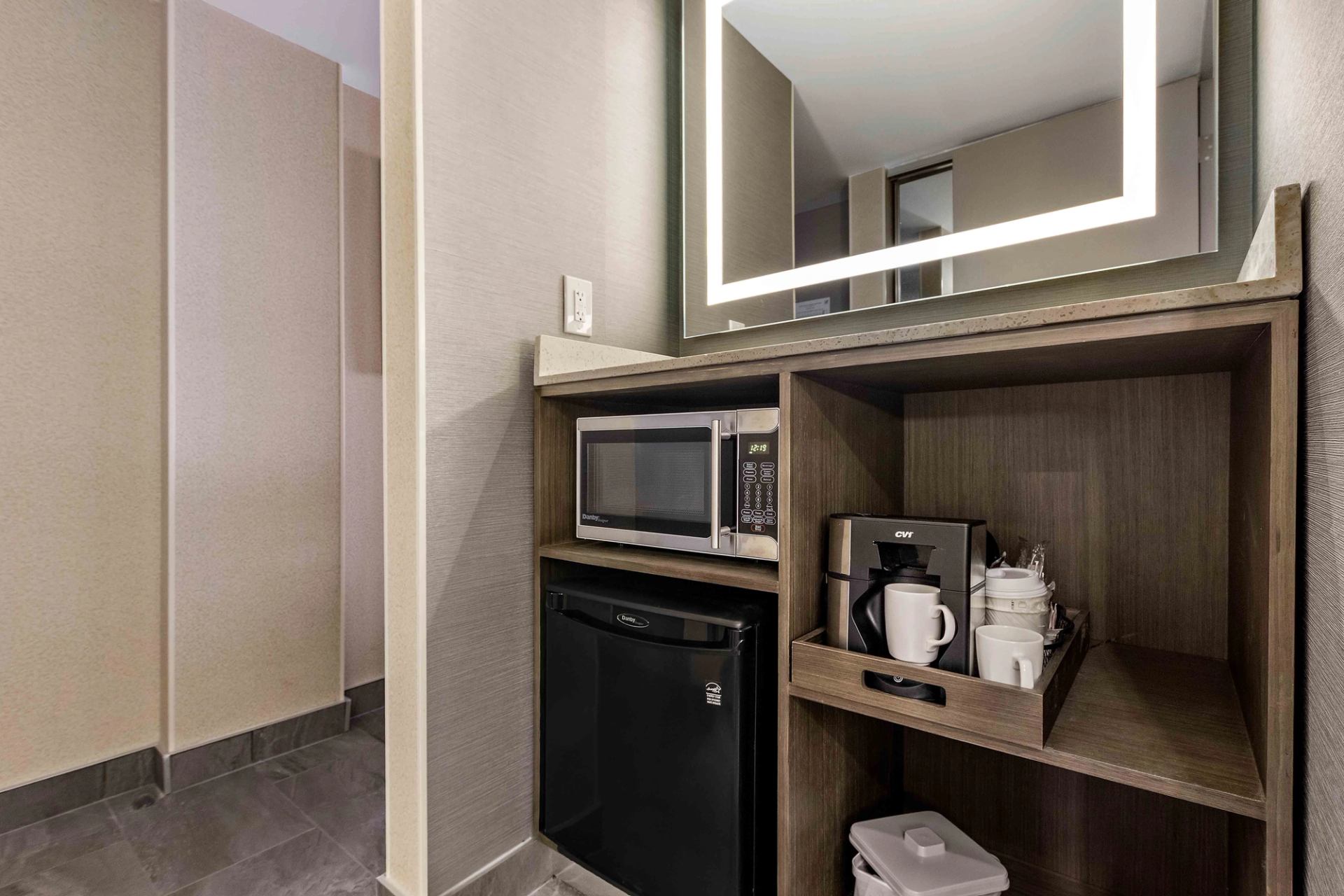 Hotel room kitchenette with microwave, mini fridge, coffee maker and cups on shelves.