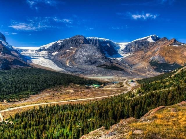 Mountain valley with glaciers, winding road, forested slopes and clear blue sky.