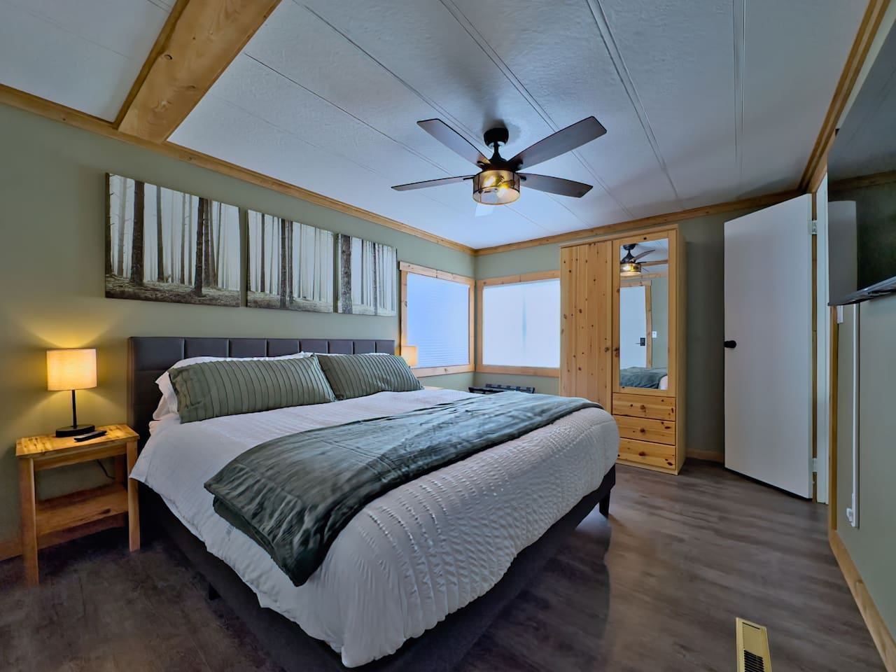 A bedroom with a king-sized bed, sage green walls, forest art, and a wooden wardrobe with a mirror.