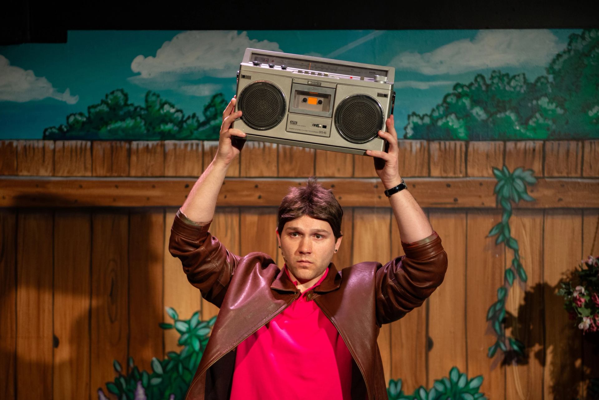 Performer in a brown jacket holds a cassette player overhead onstage.