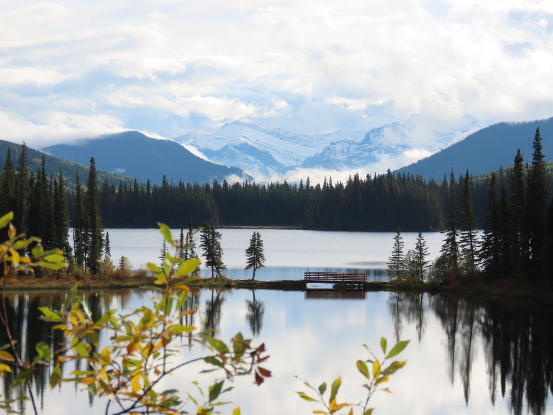 Mountain lake with forested shoreline, wooden dock and distant snow-covered peaks reflected.