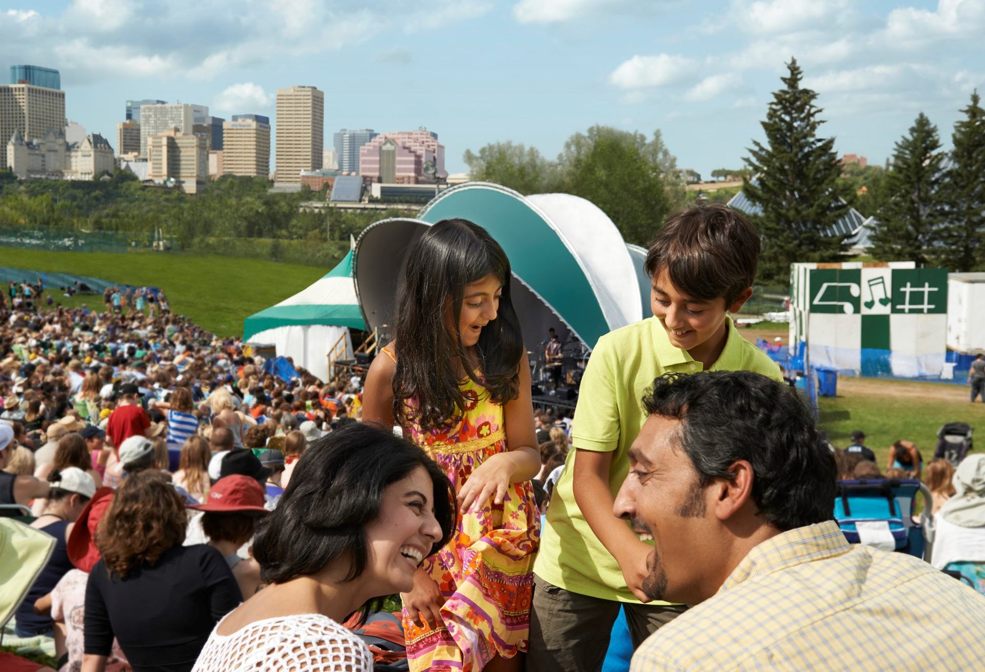 Thousands seated on grassy hill facing a stage with tents and trees in bright daylight