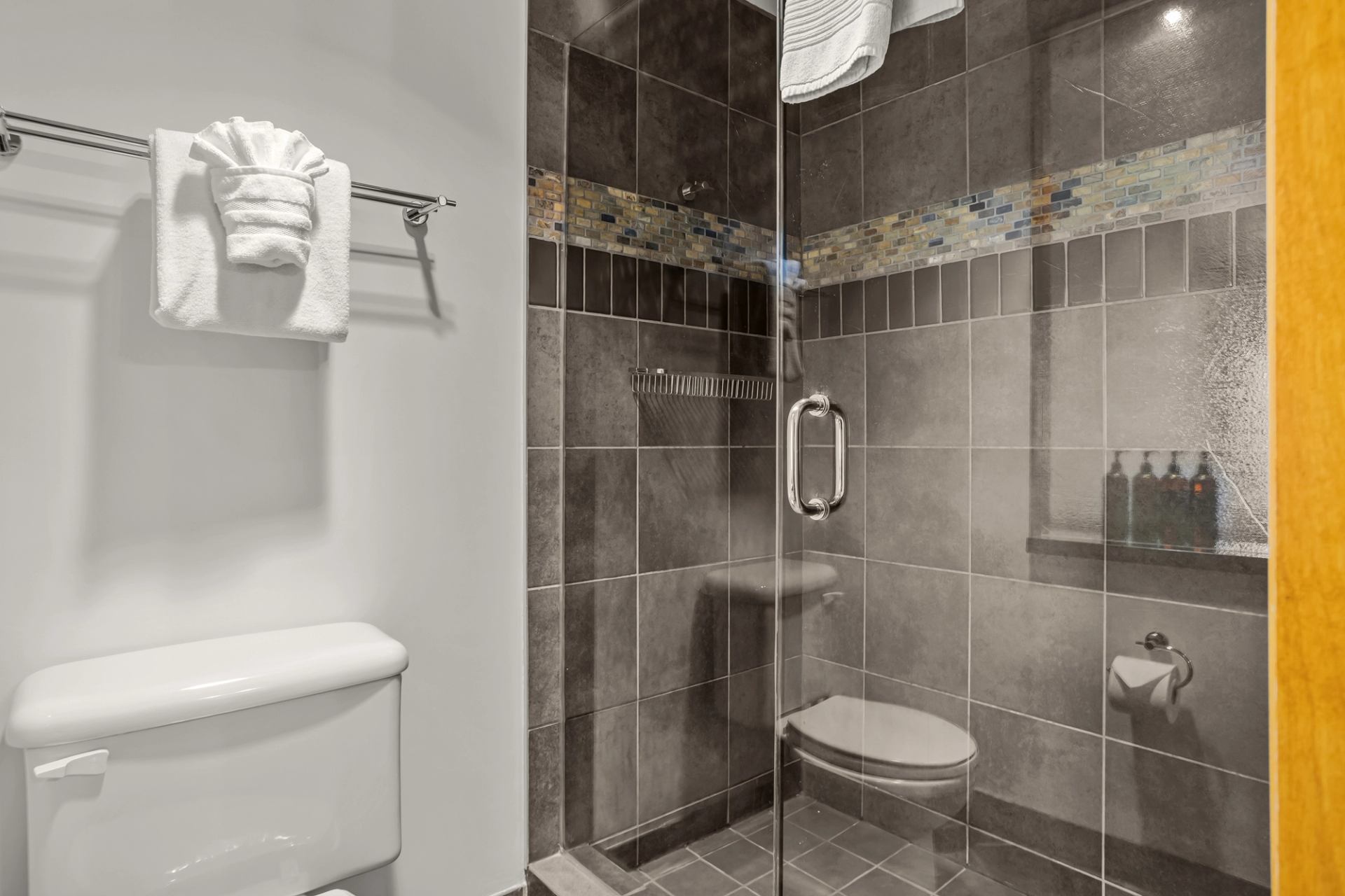 A tiled bathroom features a glass-enclosed walk-in shower, neatly folded towels, and built-in shelving with bath amenities.