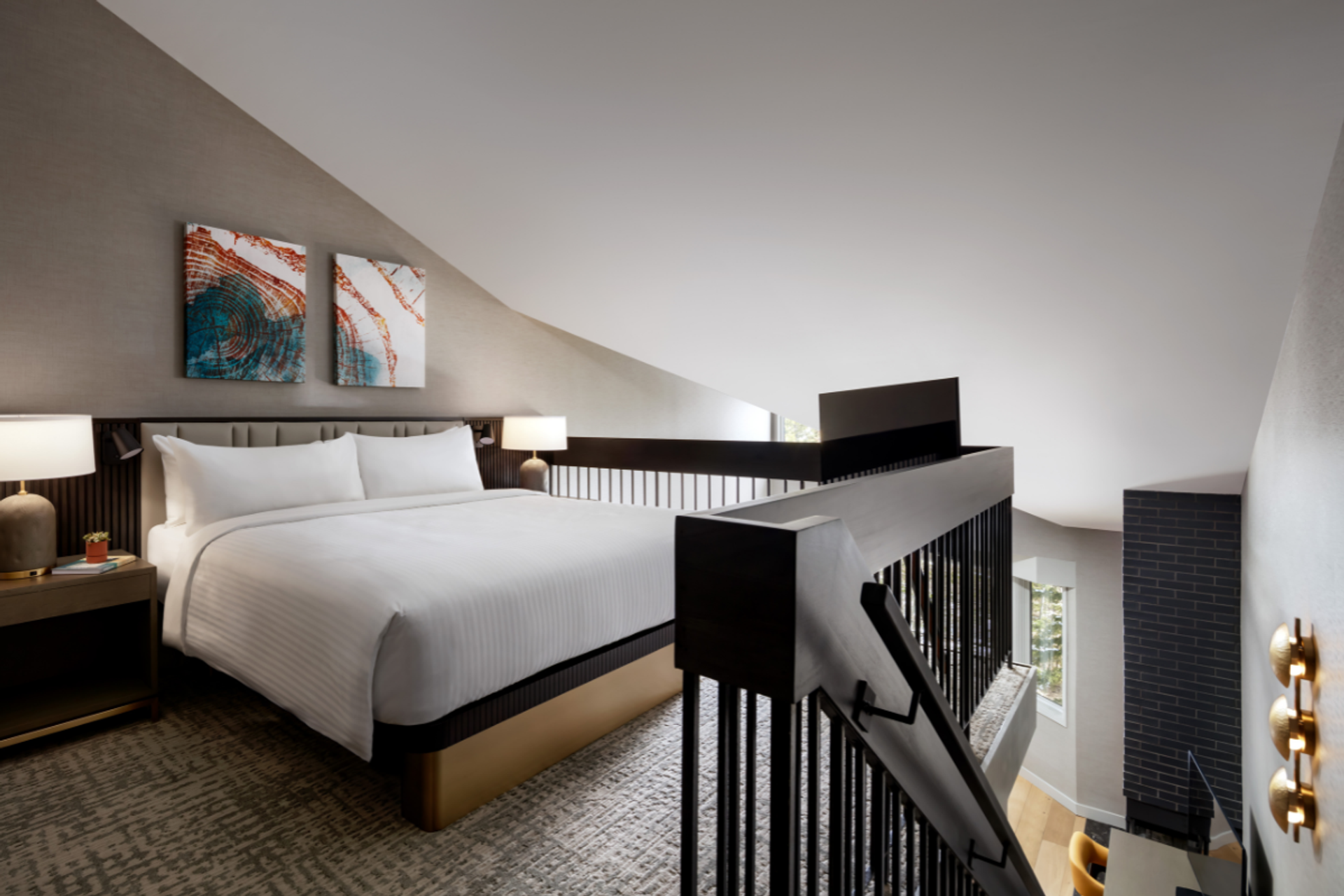 Loft bedroom with king bed, abstract artwork, lamps, and railing overlooking workspace.
