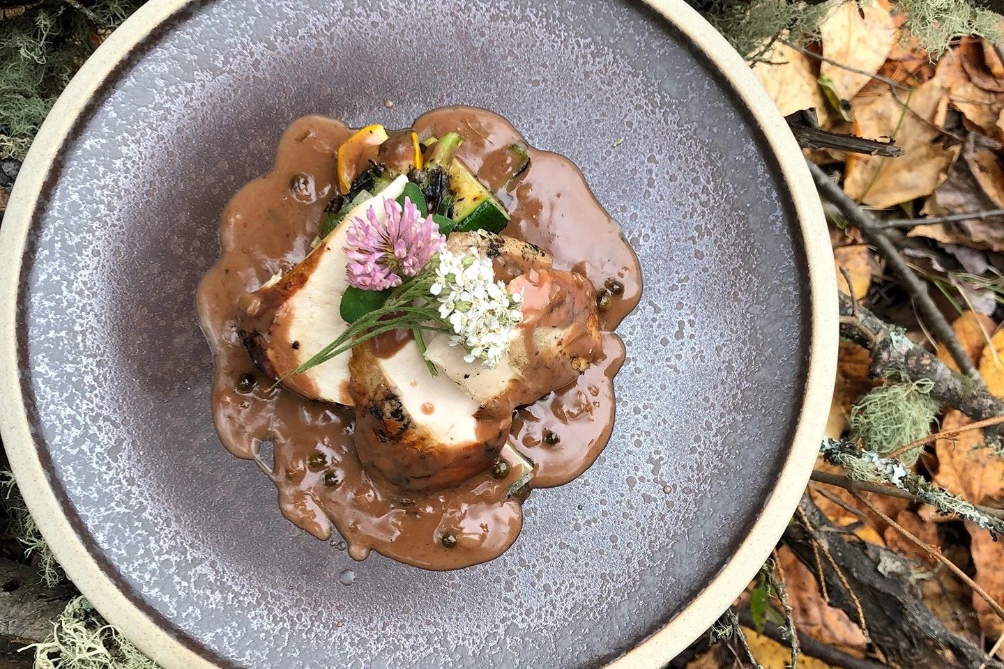 Plated roasted meat with brown sauce, grilled vegetables and edible flowers on grey dish.