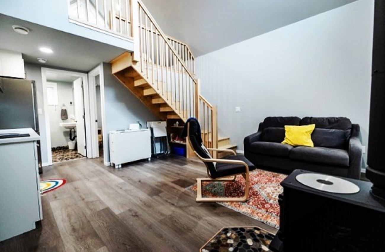 Living room with sofa, chair, loft staircase, and kitchen area in a compact bungalow.