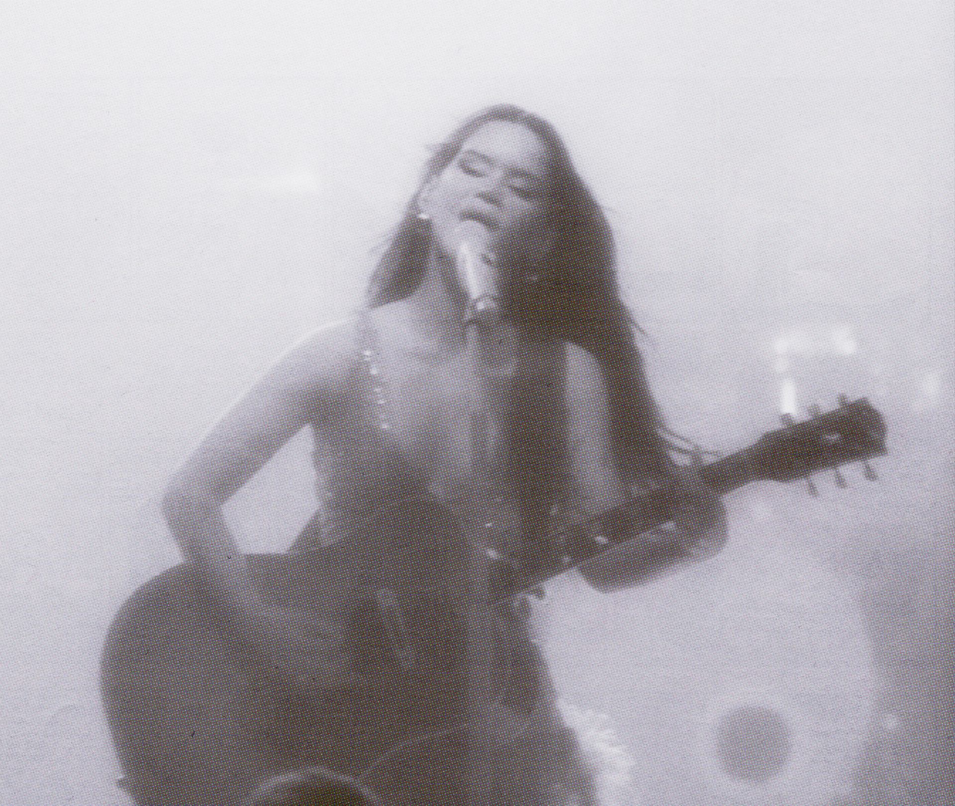 Singer with long hair plays acoustic guitar and sings into microphone on hazy stage.