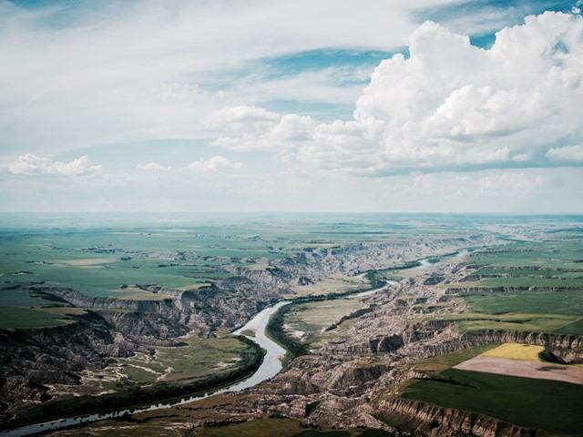 Winding river through rocky valley, farmland fields and distant plains under cloudy sky.