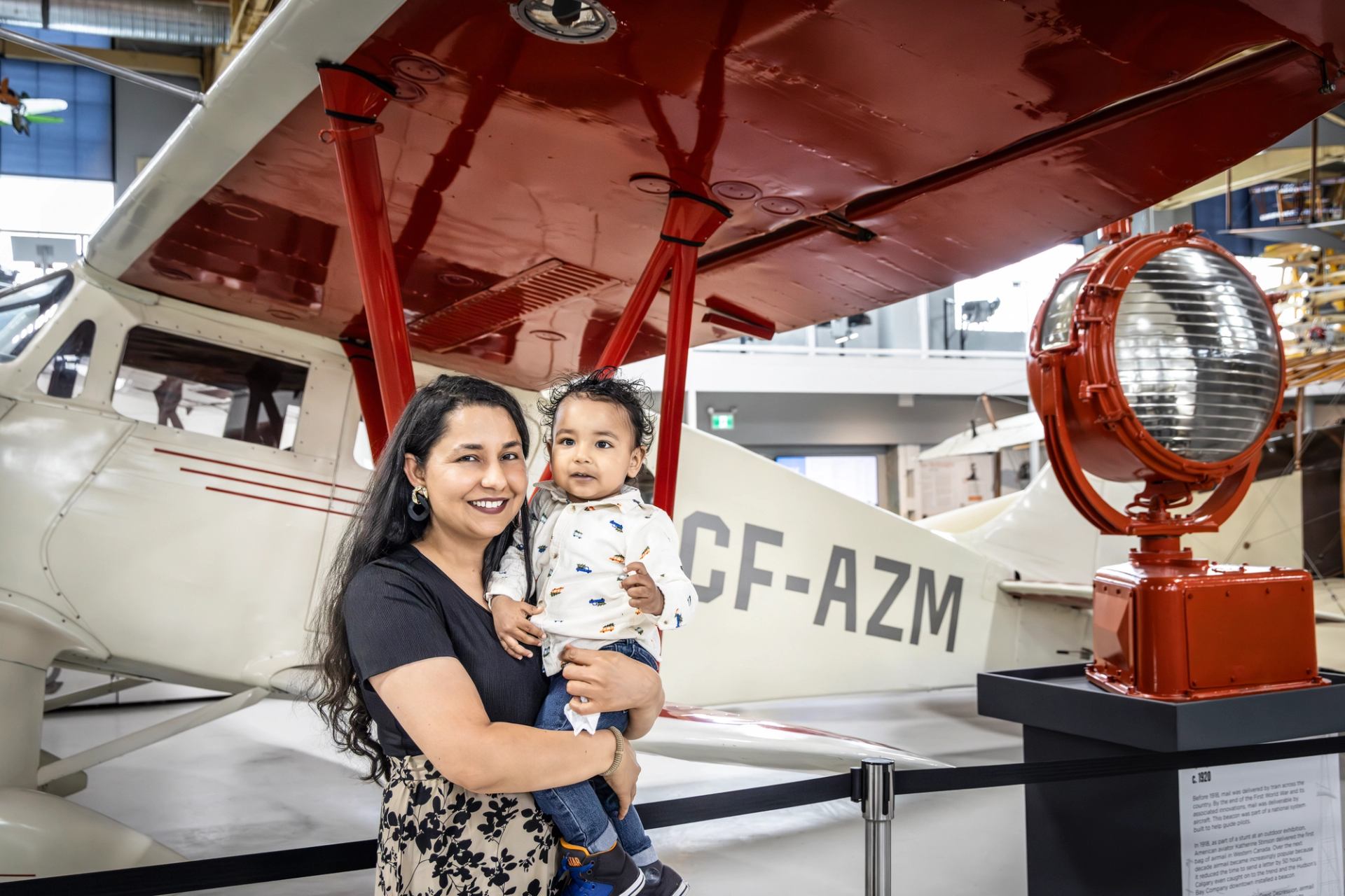A woman holding a baby smiles in front of a vintage airplane and a large red beacon light inside a museum.