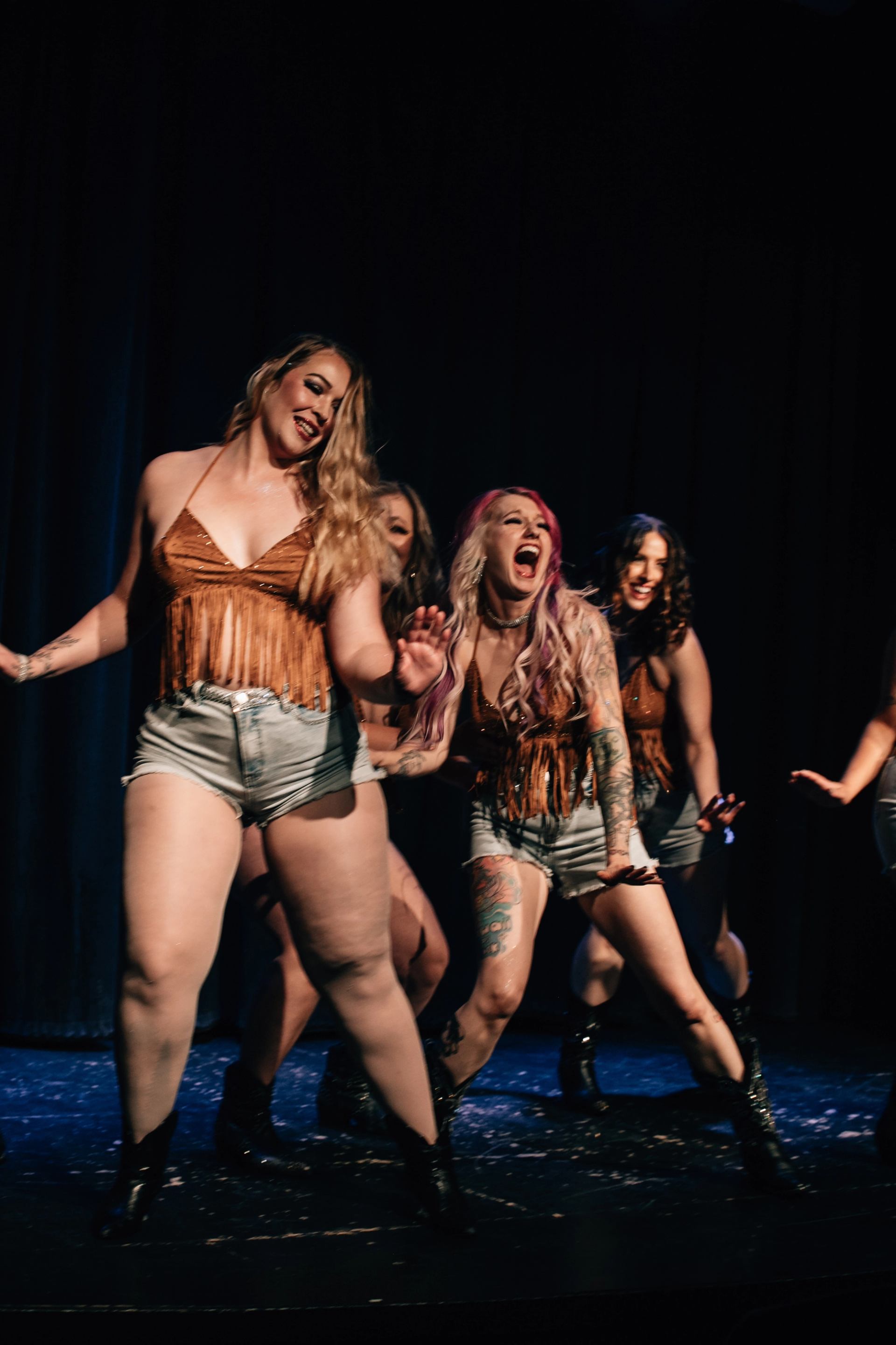 A group of women in fringed tops, denim shorts, and cowboy boots dance on a dark stage, smiling and laughing.