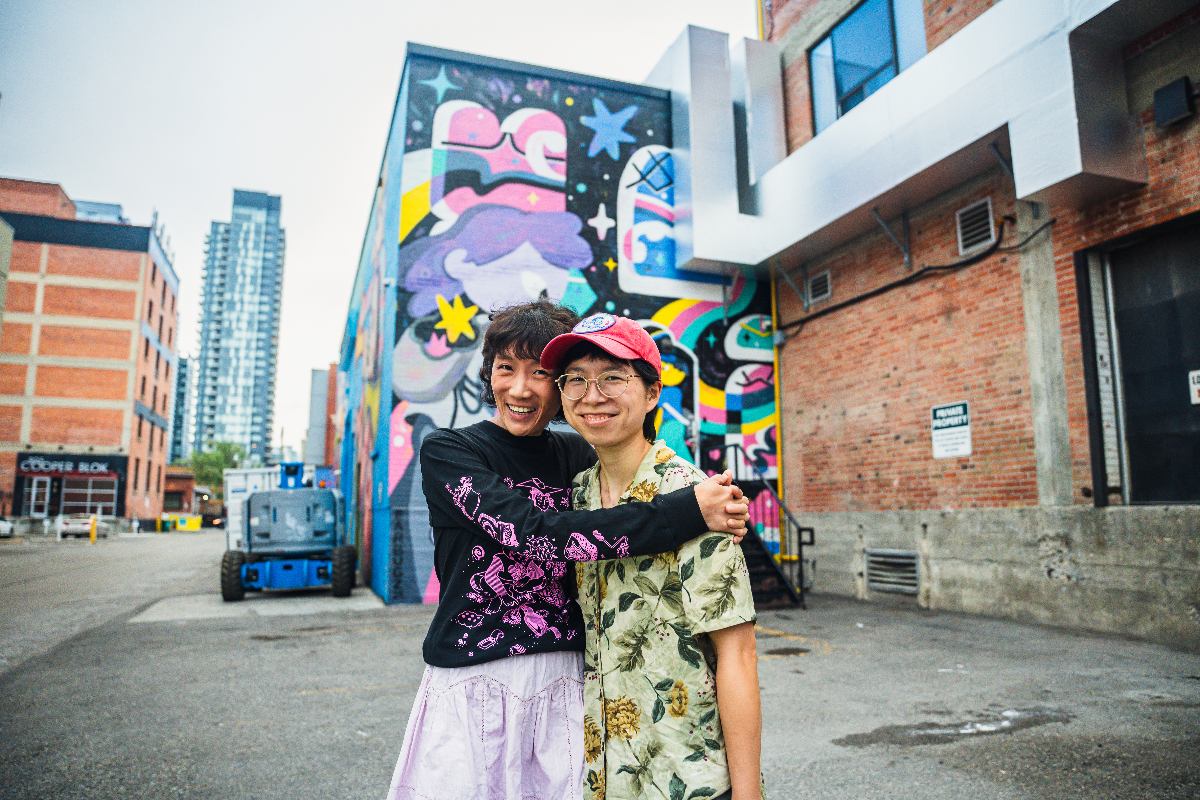 Artist Collective Earbugz (Michelle Ku and Wenting Li) embrace and smile in front of their mural titled meeting space.