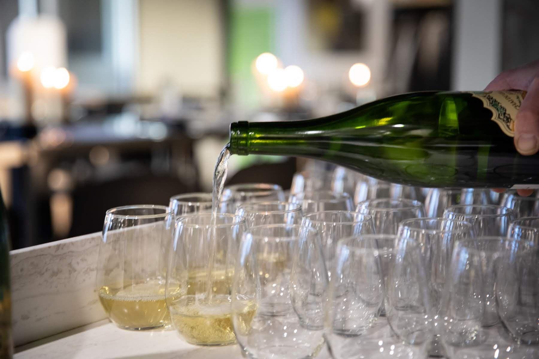 Green wine bottle pours into clear glasses on a table in a restaurant setting.