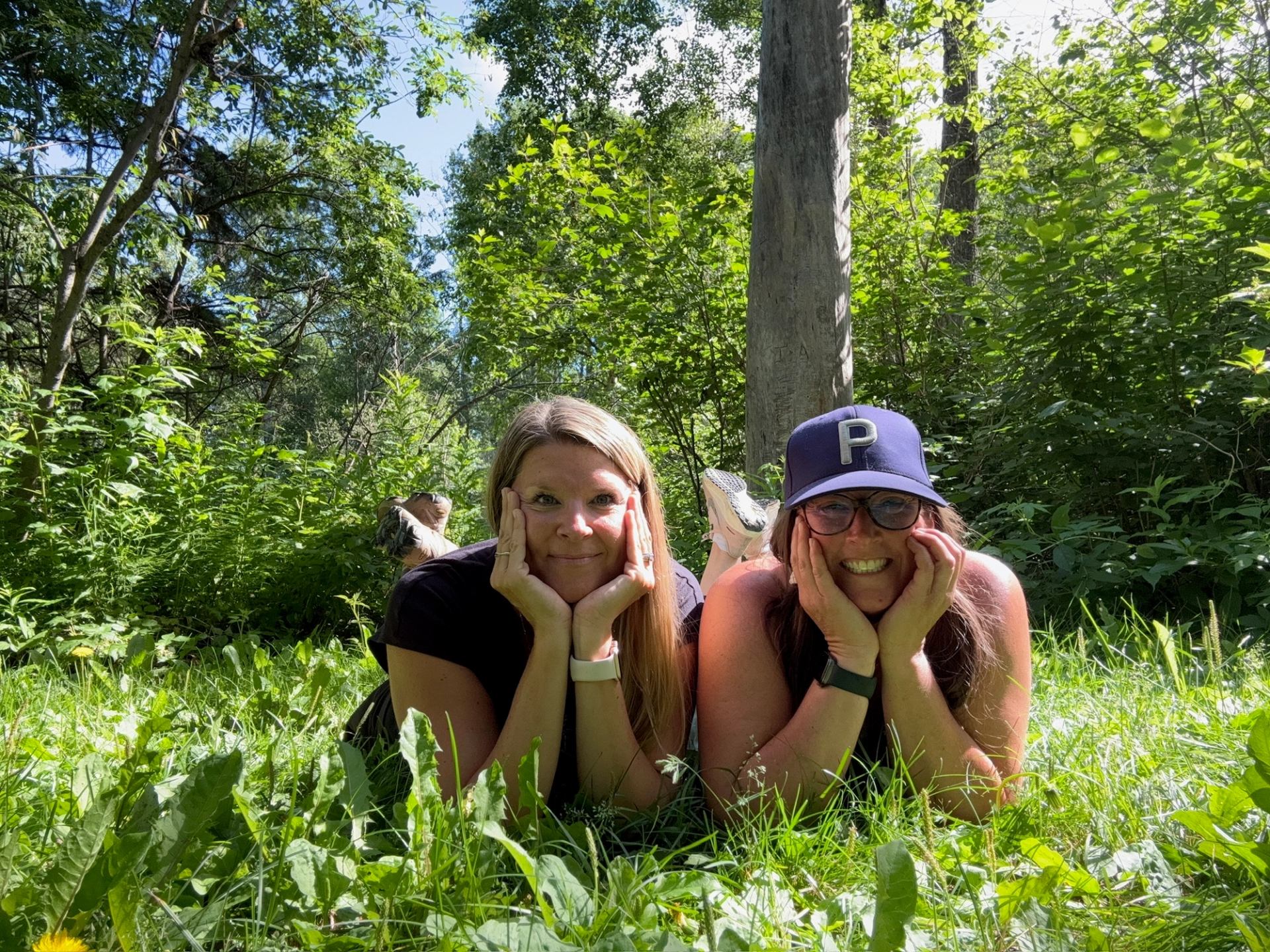Two people lying on grass in a sunny forest clearing during a hike break.